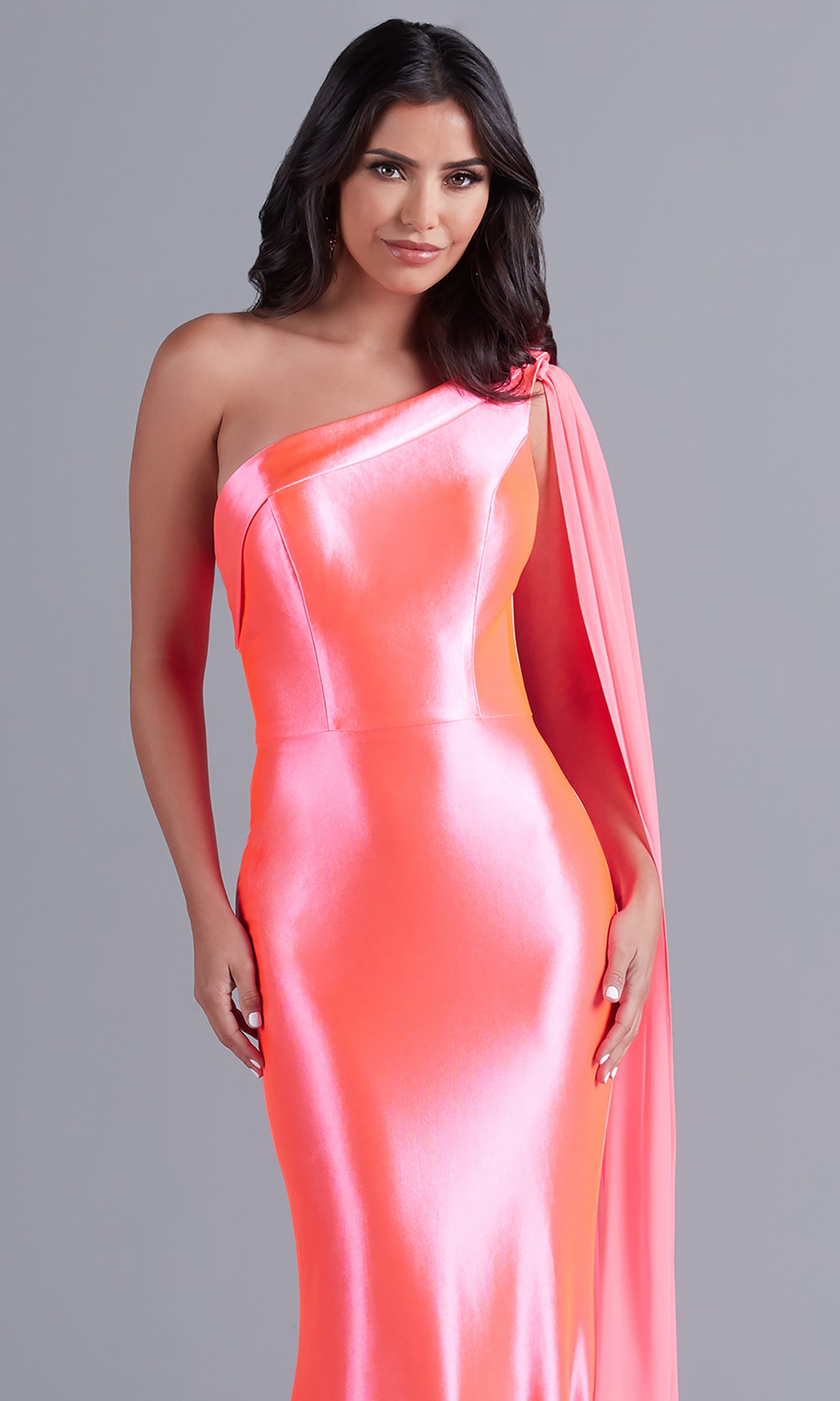 A woman models the PromGirl One-Shoulder-Sash Neon Long Prom Dress, featuring a shiny coral-pink hue and draped one-shoulder sleeve, set against a plain gray background.